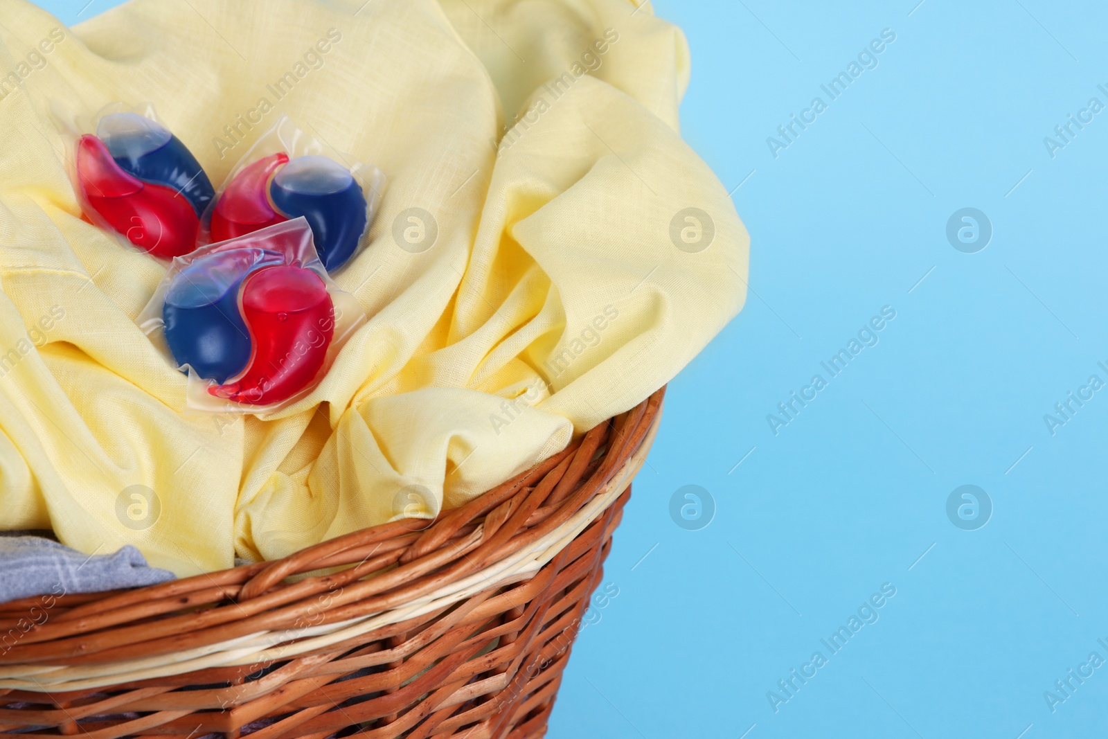 Photo of Detergent capsules and laundry in basket on light blue background, closeup