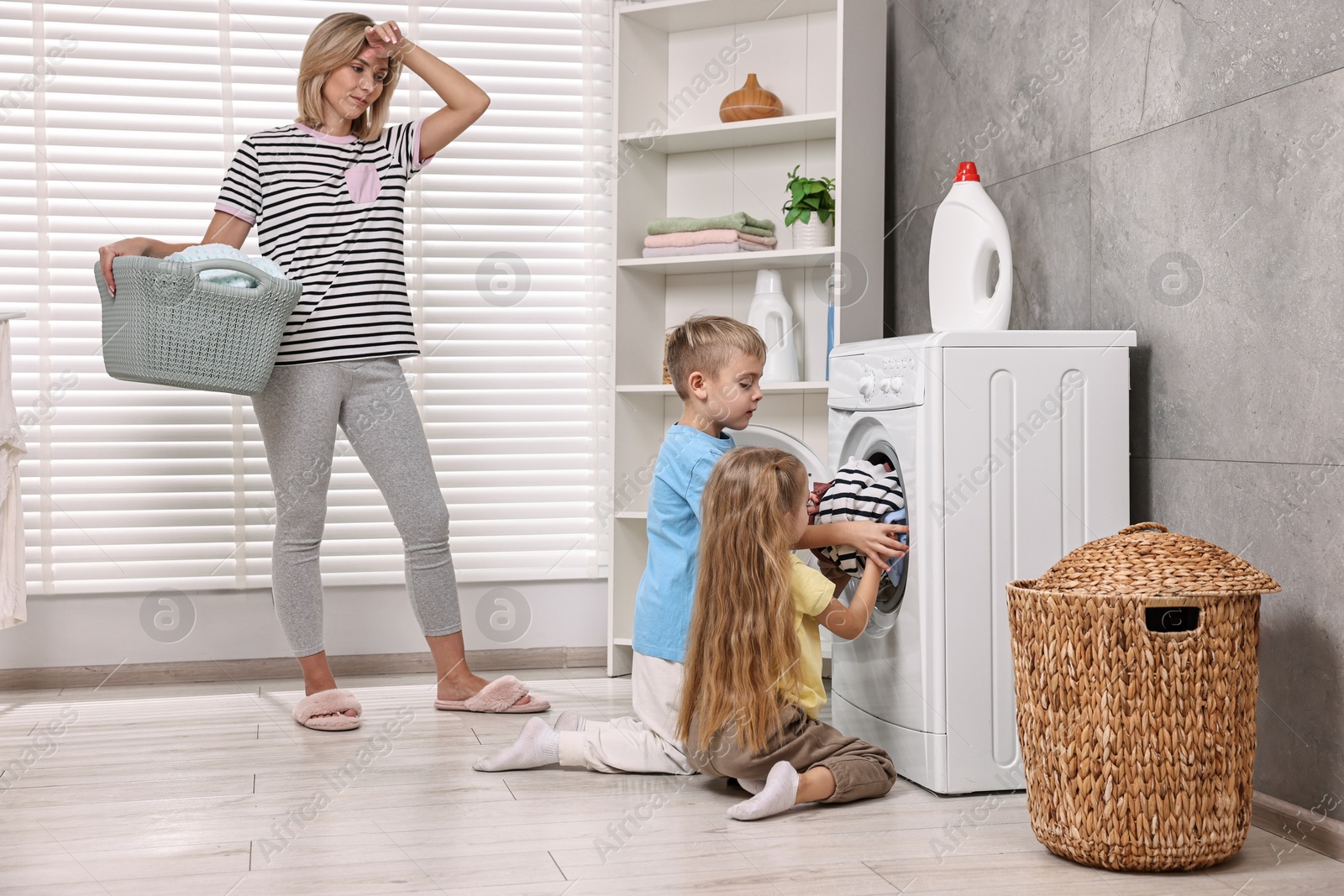 Photo of Tired housewife and her kids doing laundry together in bathroom