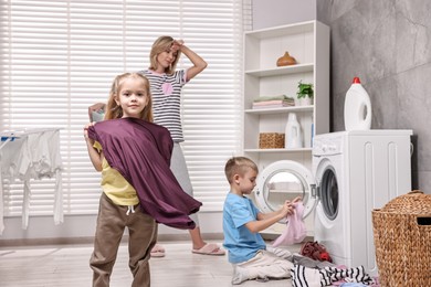 Photo of Tired housewife and her kids doing laundry together in bathroom