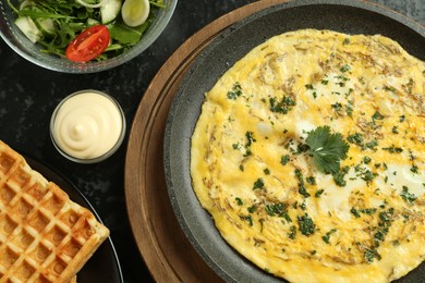 Photo of Tasty brunch. Delicious omelette, salad and waffles served on black textured table, flat lay