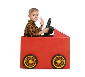 Photo of Little boy waving while driving car made of cardboard on white background