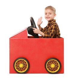 Photo of Little boy waving while driving car made of cardboard on white background