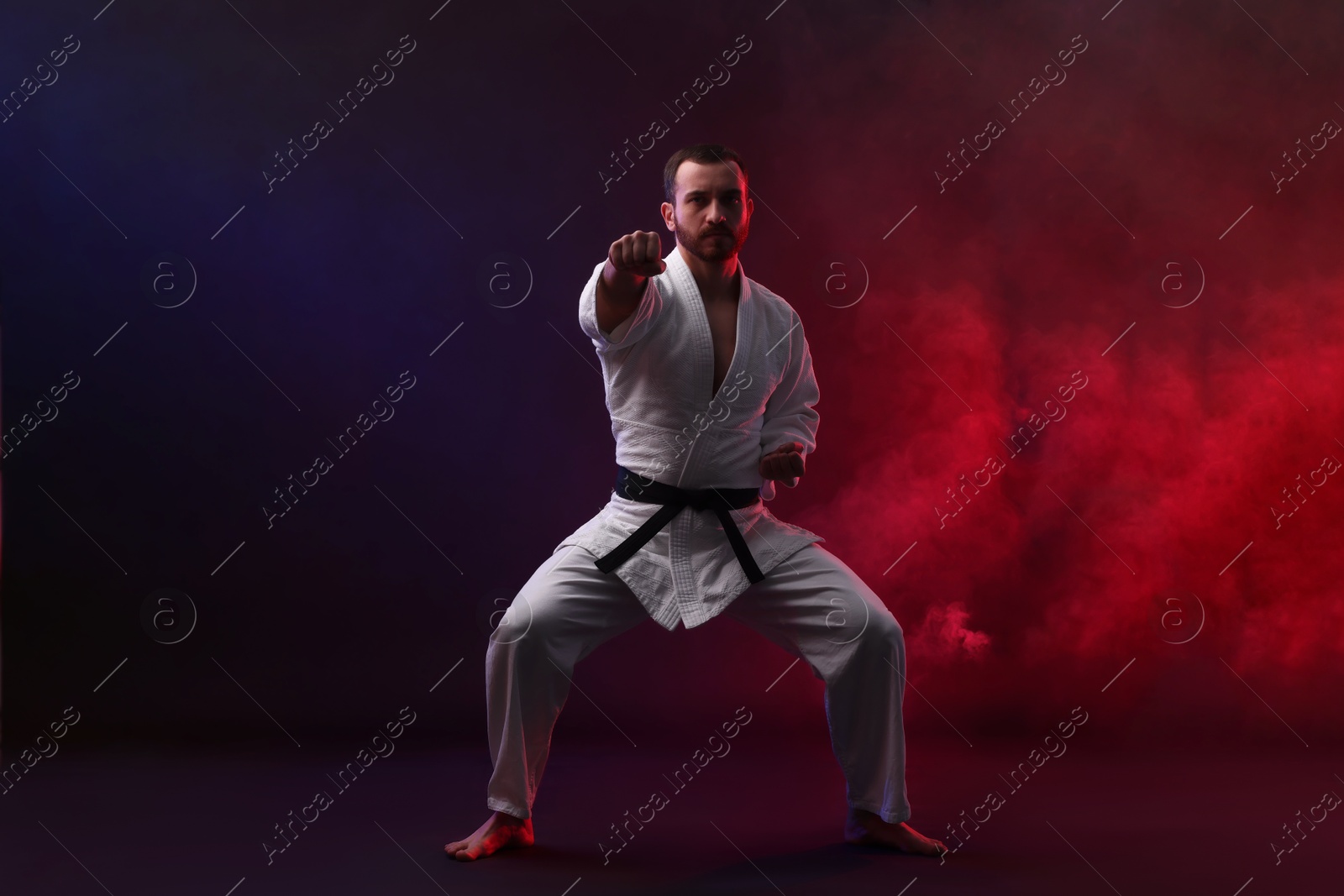 Photo of Man wearing uniform practicing karate in color lights and smoke against black background