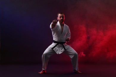 Photo of Man wearing uniform practicing karate in color lights and smoke against black background