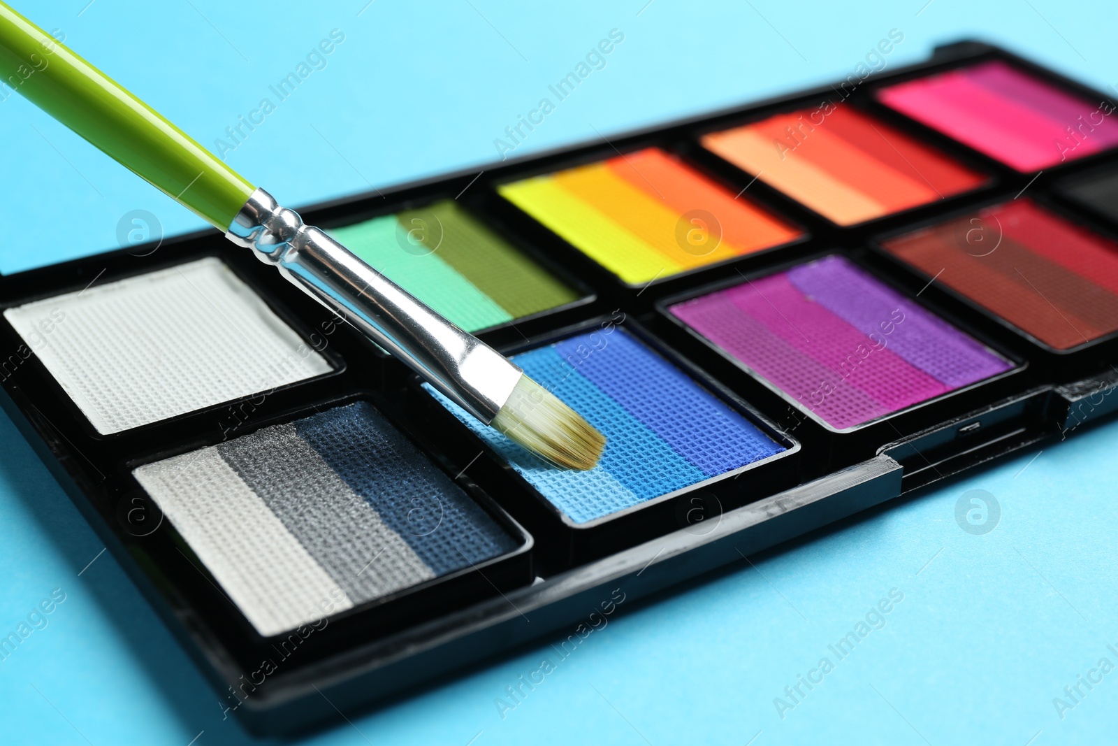 Photo of Taking face paint with brush from palette on light blue background, closeup