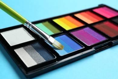 Photo of Taking face paint with brush from palette on light blue background, closeup