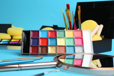 Photo of Bright face paints, brushes, mirror and sponges on light blue background, closeup