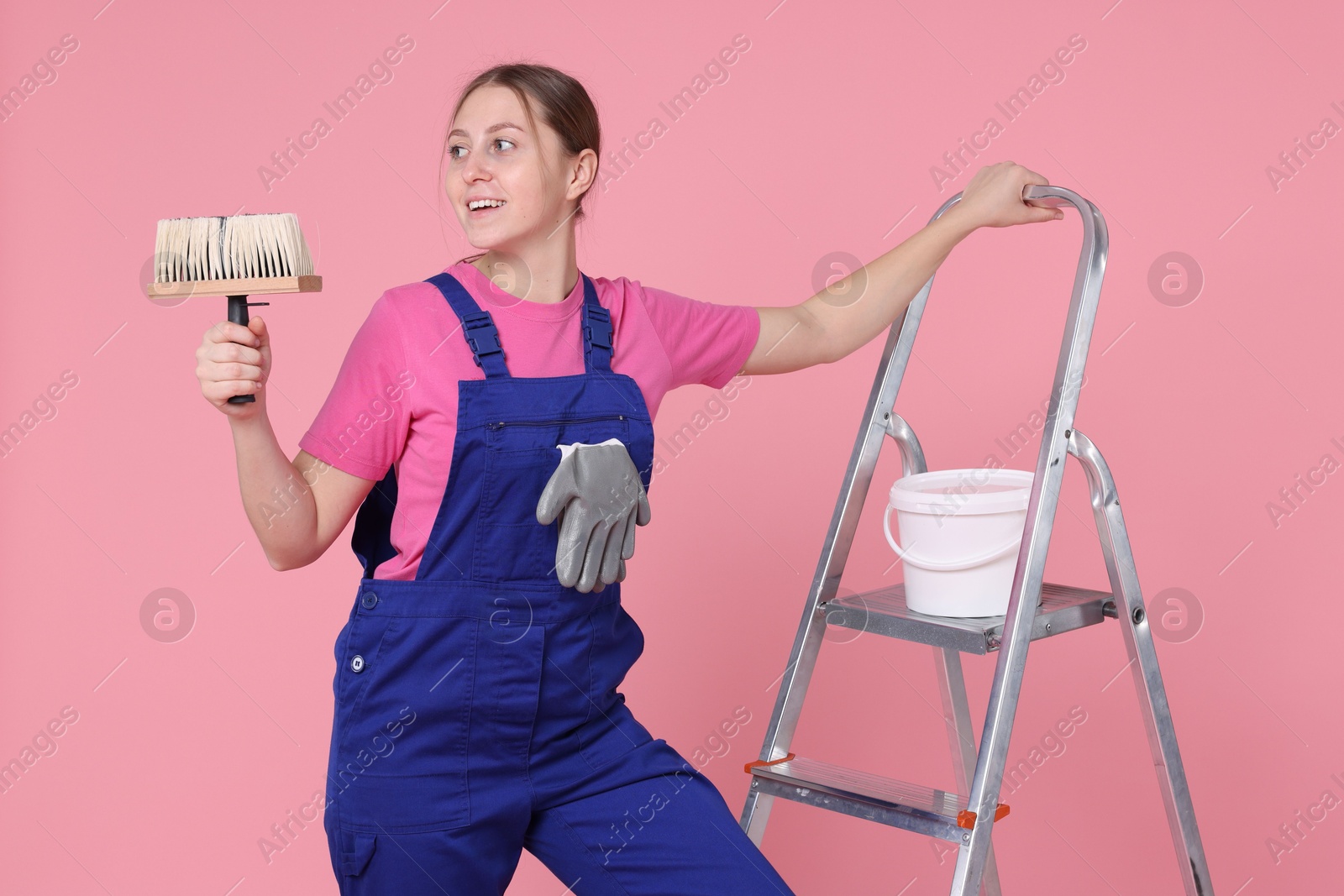 Photo of Professional painter with brush, bucket of paint and ladder on light pink background