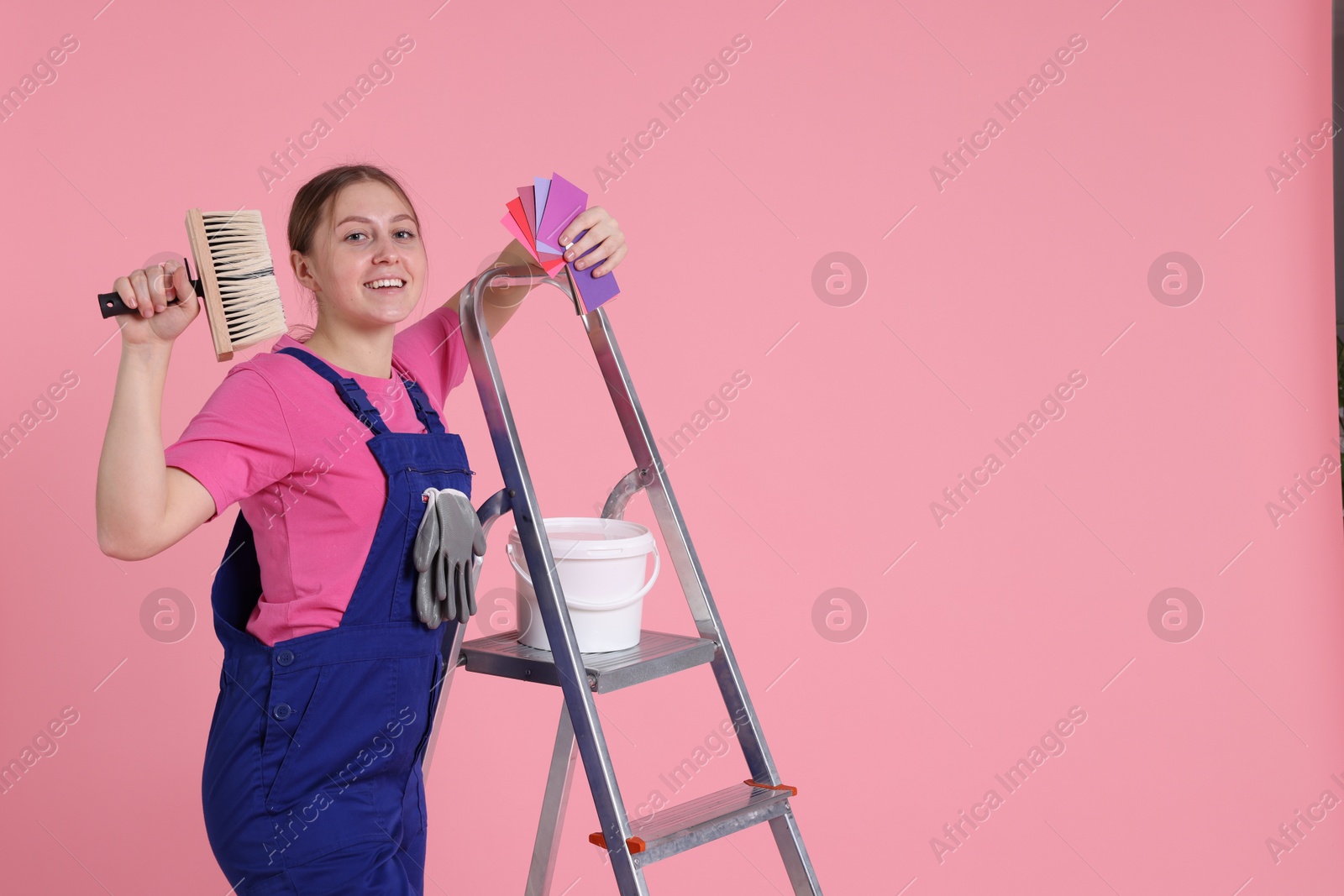 Photo of Professional painter with brush, color samples, bucket of paint and ladder on light pink background. Space for text