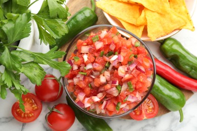 Delicious salsa in bowl, nacho chips and ingredients on white marble table, flat lay