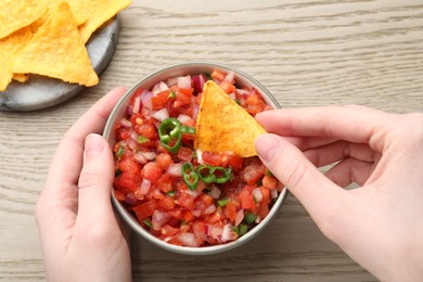 Photo of Woman dipping nacho chip into delicious salsa sauce at wooden table, top view