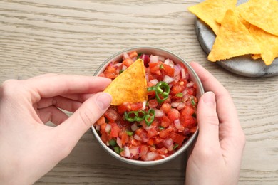 Woman dipping nacho chip into delicious salsa sauce at wooden table, top view