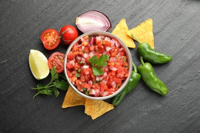 Delicious salsa in bowl, nacho chips and ingredients on grey table, flat lay