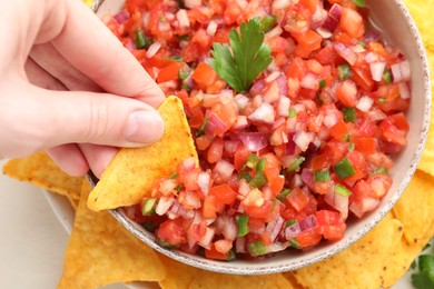 Photo of Woman dipping nacho chip into delicious salsa sauce at table, top view