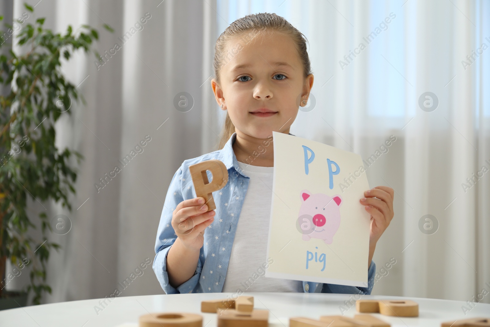 Photo of Learning alphabet. Little girl with wooden letter P and picture at white table indoors