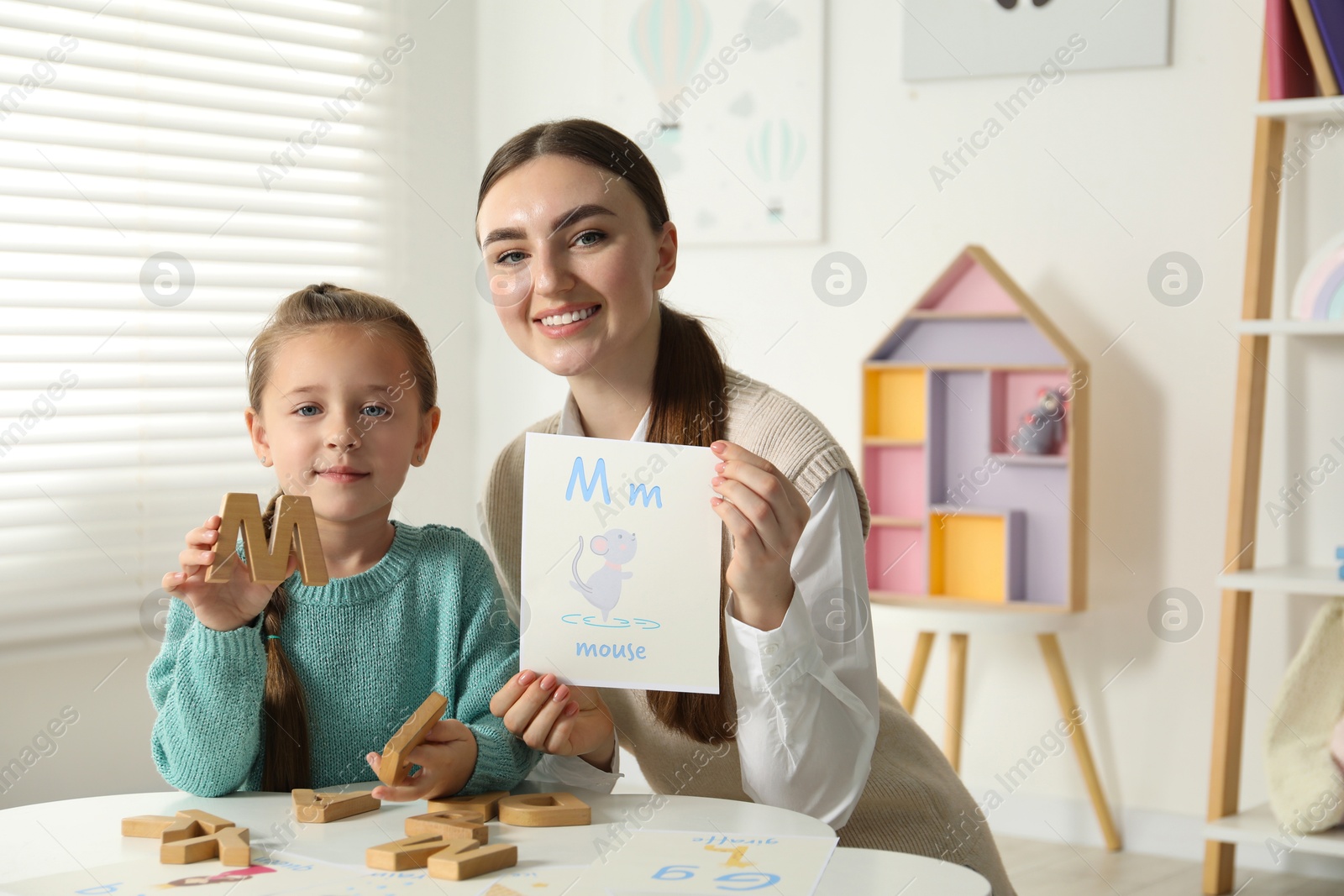 Photo of Speech therapist teaching little girl alphabet at white table indoors