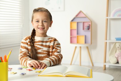 Photo of Little girl learning alphabet with magnetic letters at white table indoors