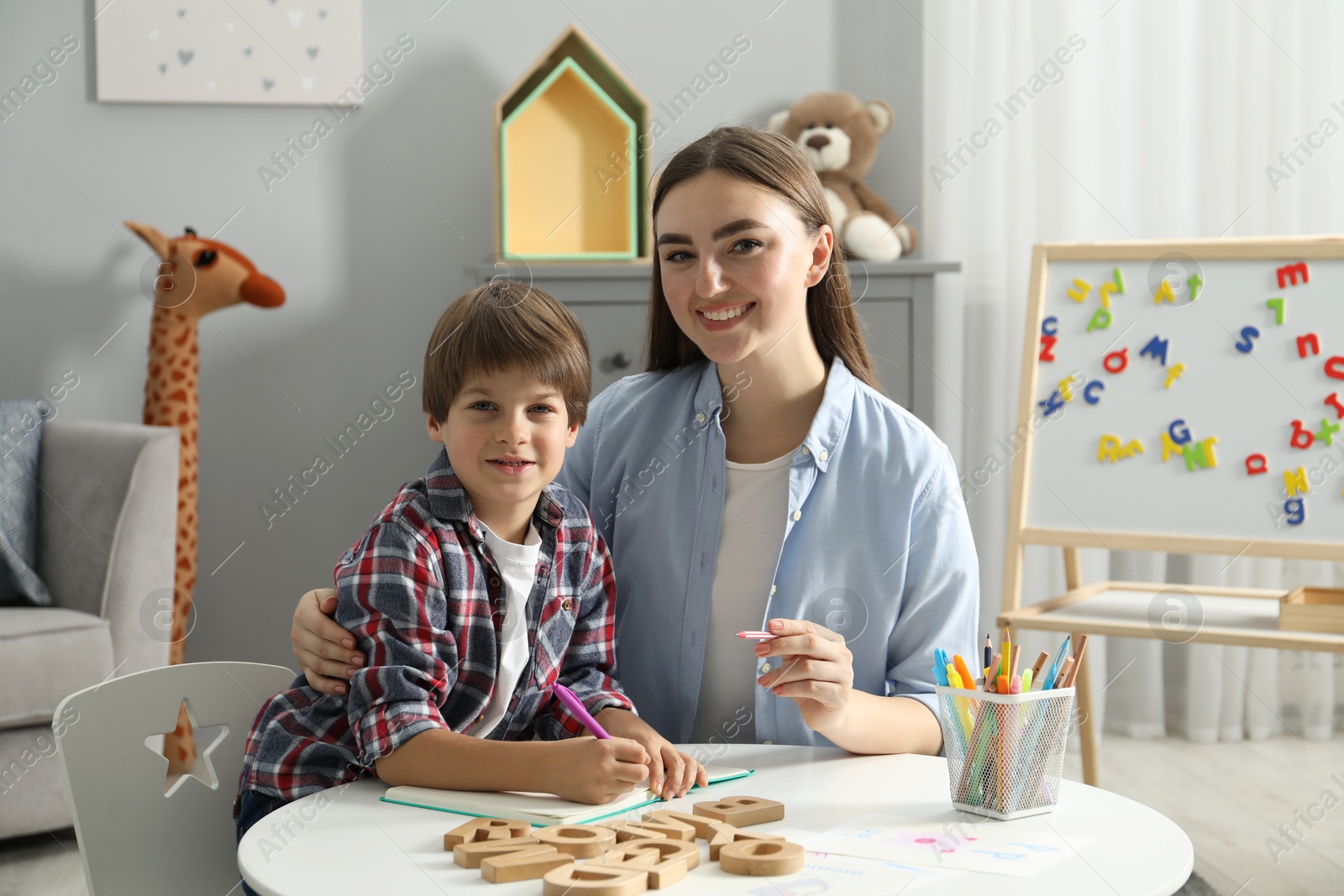 Photo of Speech therapist teaching little boy alphabet at white table indoors