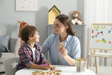 Photo of Speech therapist teaching little boy alphabet with wooden letters at white table indoors