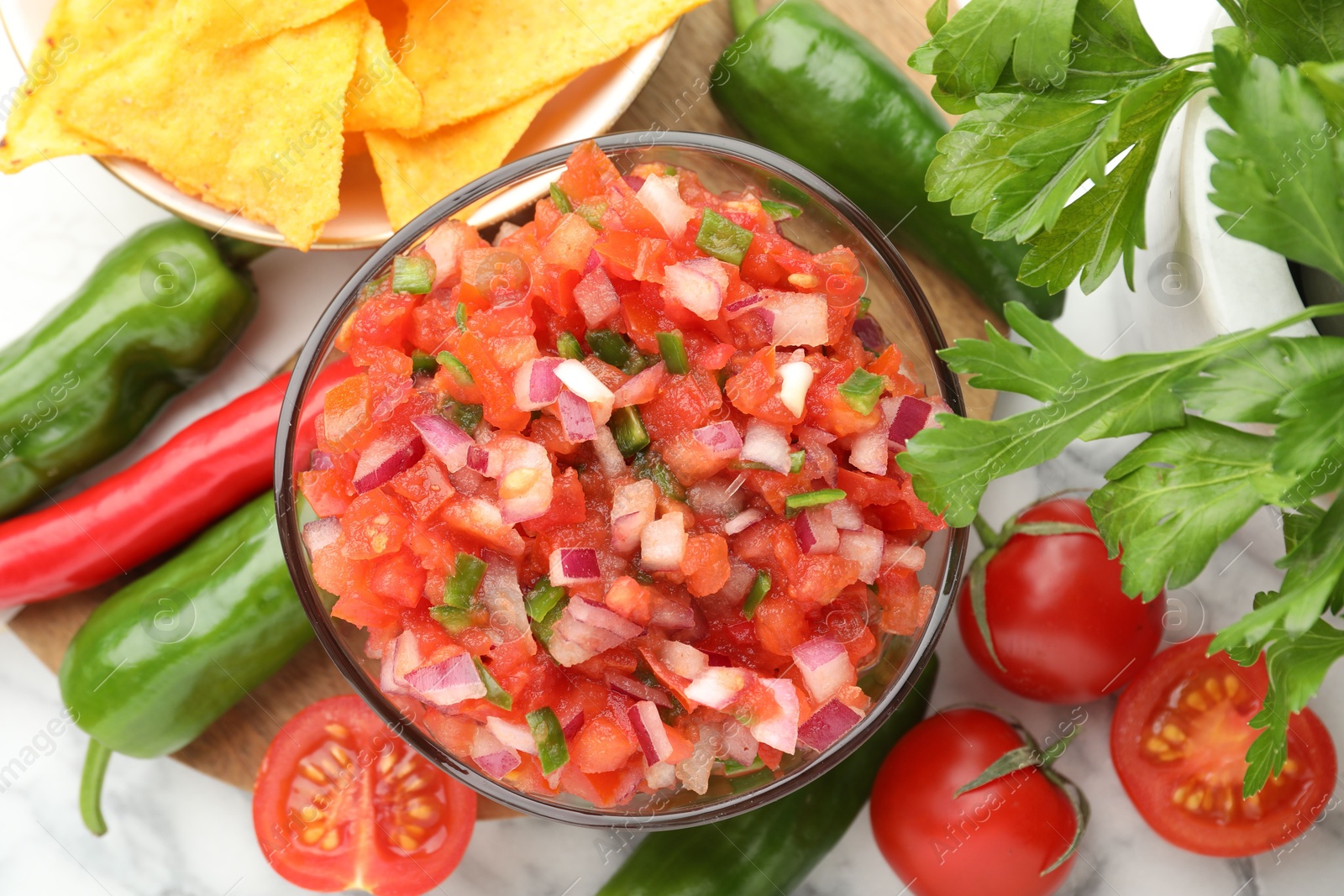 Photo of Delicious salsa in bowl, nacho chips and ingredients on white marble table, flat lay