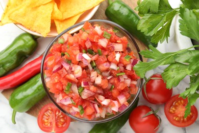 Photo of Delicious salsa in bowl, nacho chips and ingredients on white marble table, flat lay