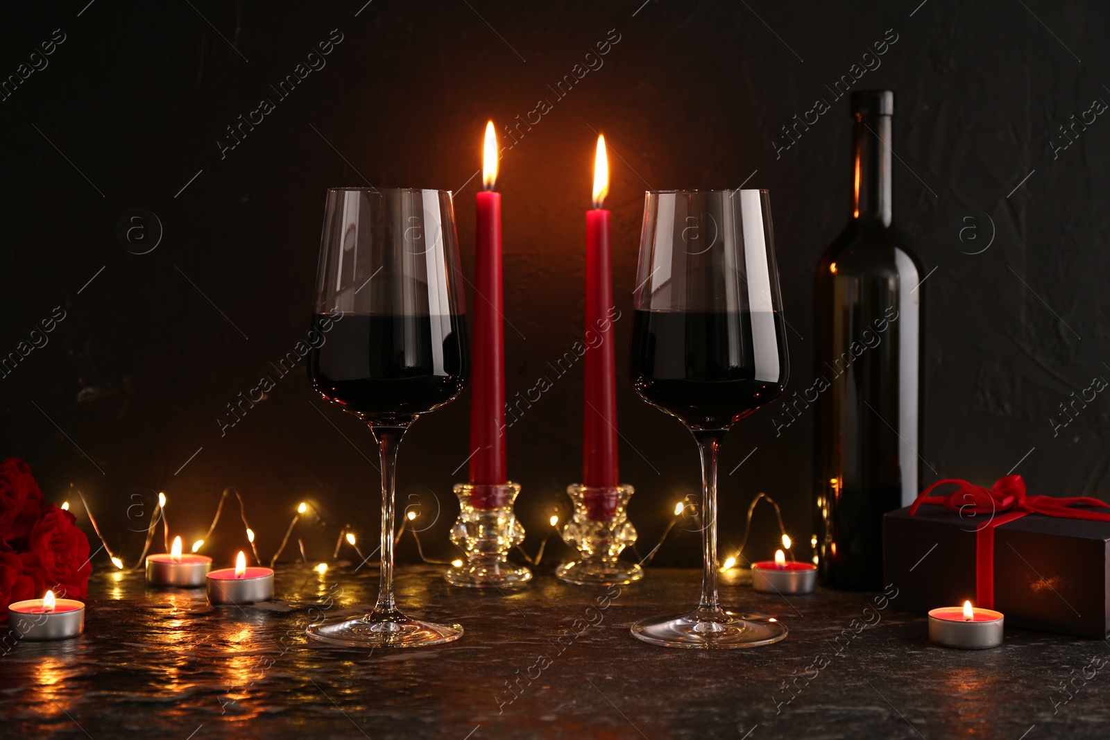 Photo of Glasses of wine, burning candles, gift box and roses on black textured table. Romantic dinner