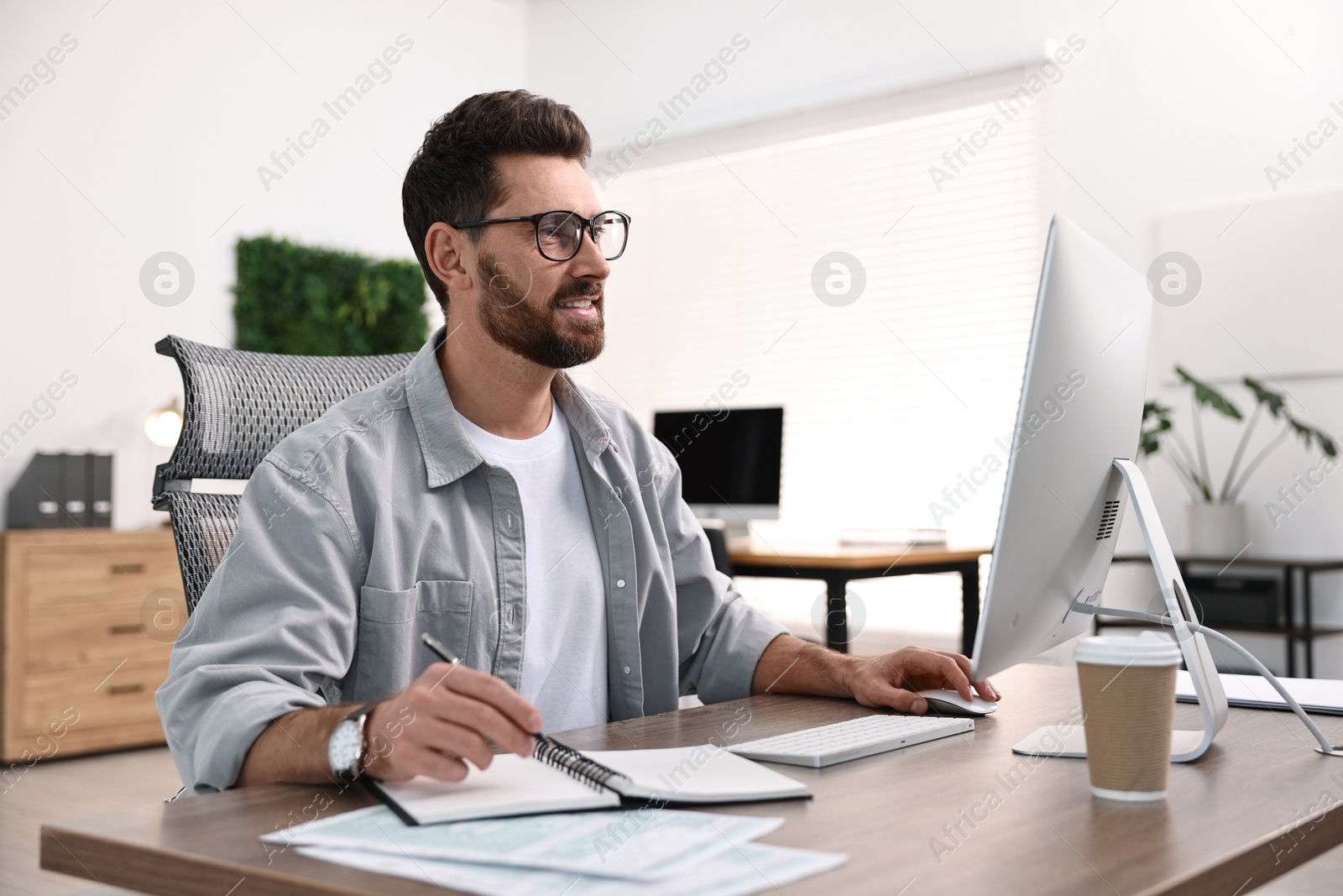 Photo of Man taking notes while working on computer at table in office