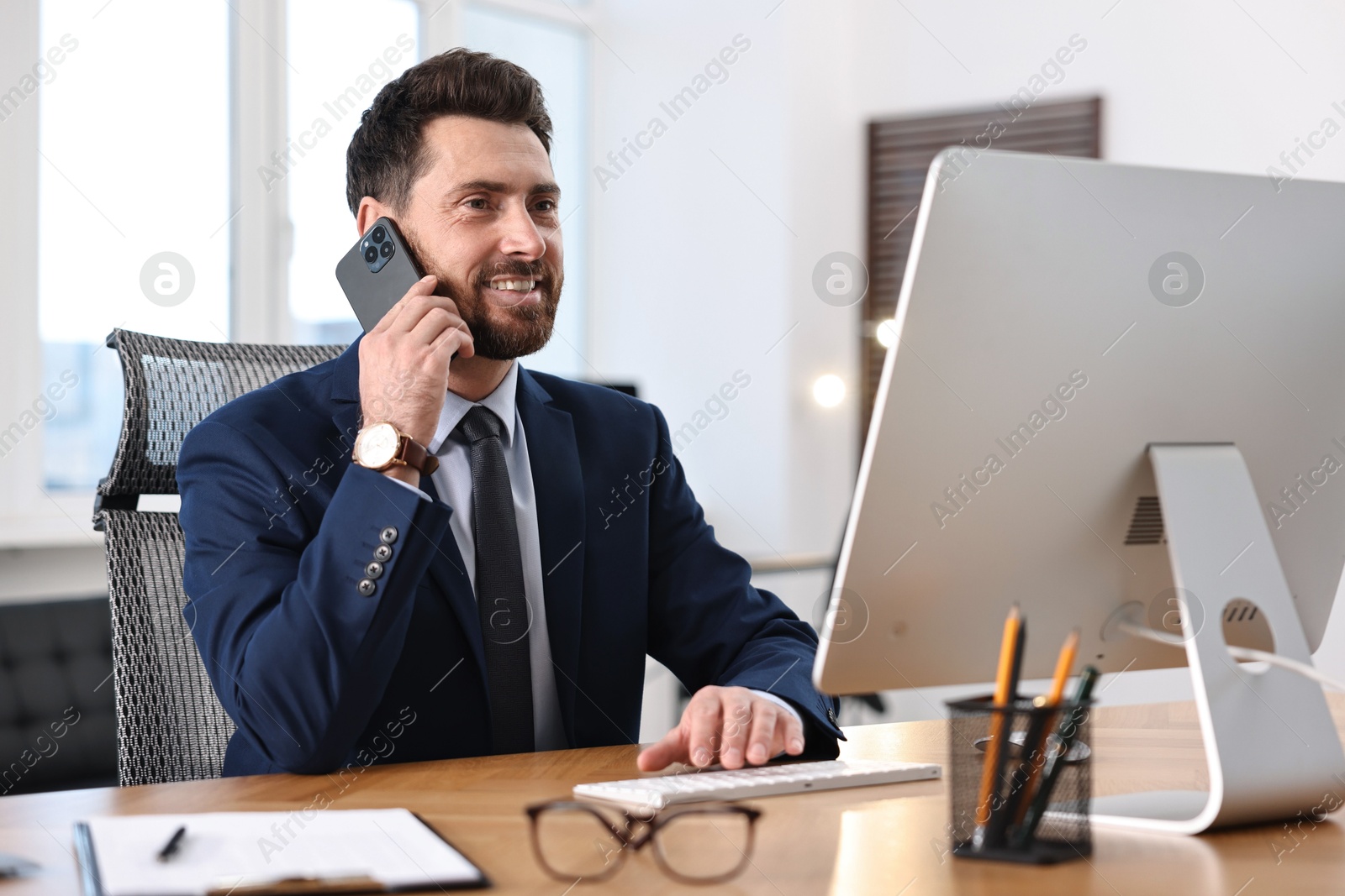 Photo of Man talking on smartphone while working with computer at table in office