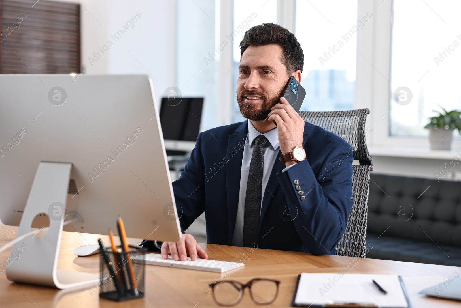 Photo of Man talking on smartphone while working with computer at table in office