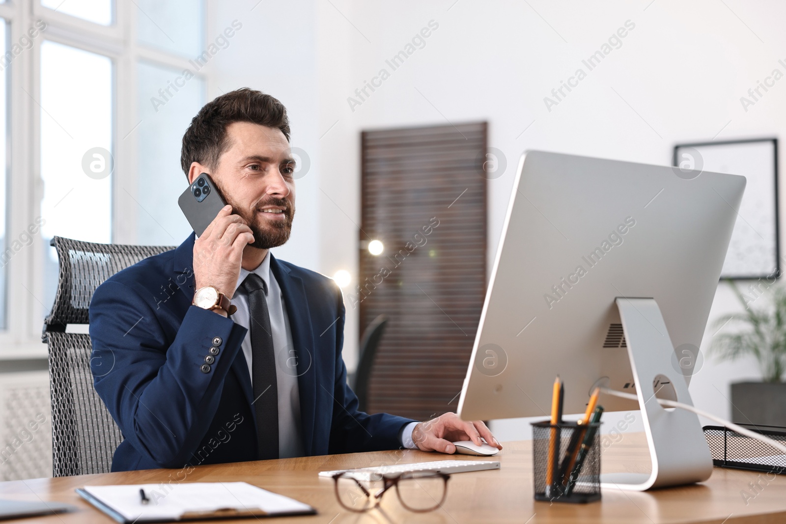 Photo of Man talking on smartphone while working with computer at table in office