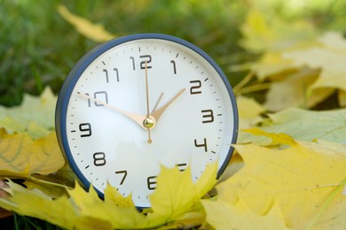 Photo of Autumn time. Alarm clock among fallen leaves on green grass, closeup