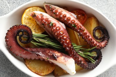 Photo of Fried octopus with vegetables, lemon and rosemary in baking dish on grey textured table, top view