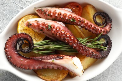 Photo of Fried octopus with vegetables, lemon and rosemary in baking dish on grey textured table, top view