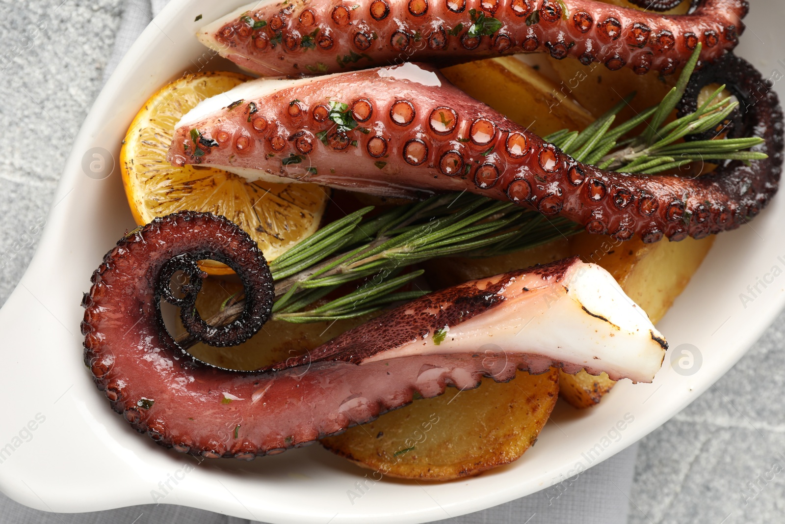 Photo of Fried octopus with vegetables, lemon and rosemary in baking dish on grey textured table, top view