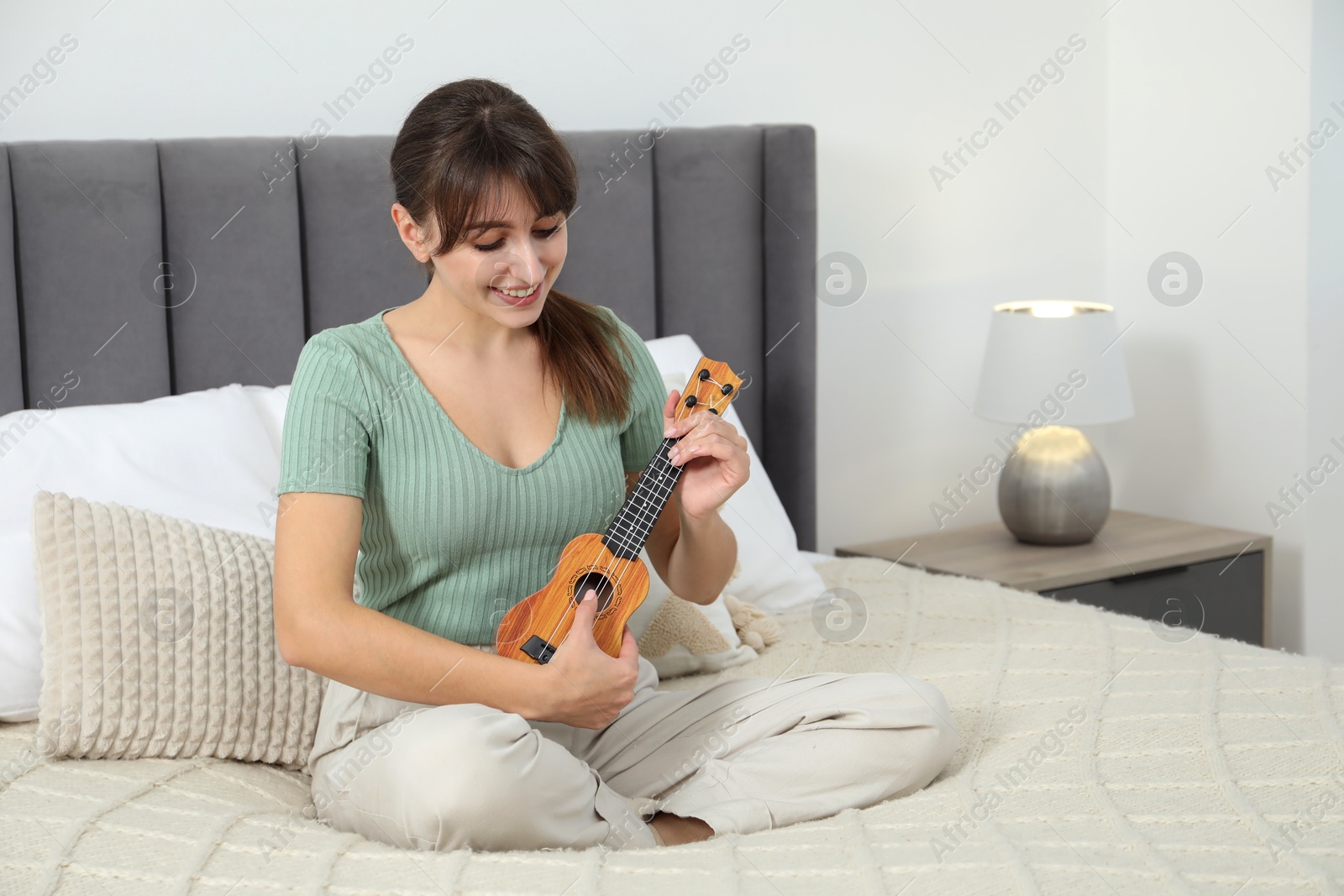 Photo of Happy woman playing ukulele on bed at home