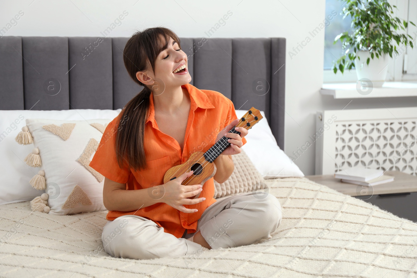 Photo of Happy woman playing ukulele on bed at home