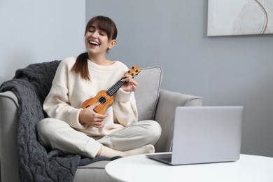 Photo of Woman learning to play ukulele with online music course in armchair at home