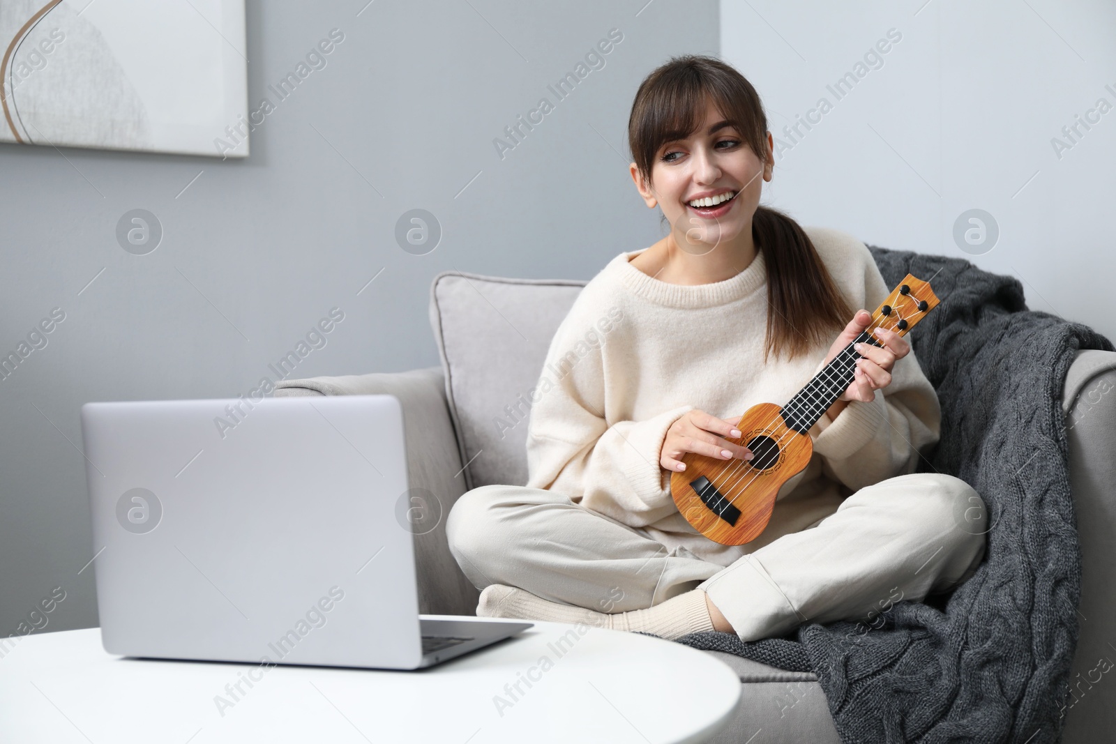 Photo of Woman learning to play ukulele with online music course in armchair at home