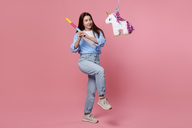 Photo of Happy woman hitting unicorn shaped pinata with stick on pink background