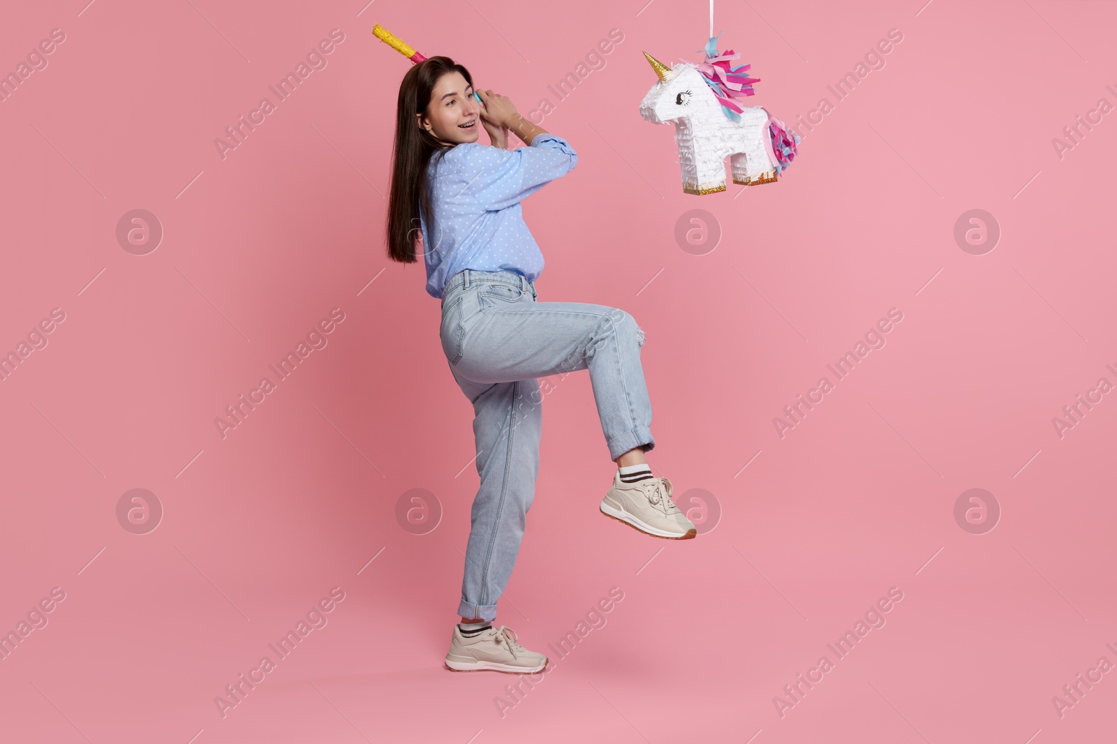 Photo of Happy woman hitting unicorn shaped pinata with stick on pink background