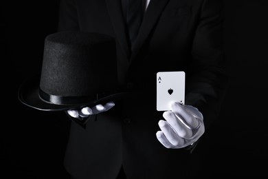 Illusionist with playing card and hat on black background, closeup