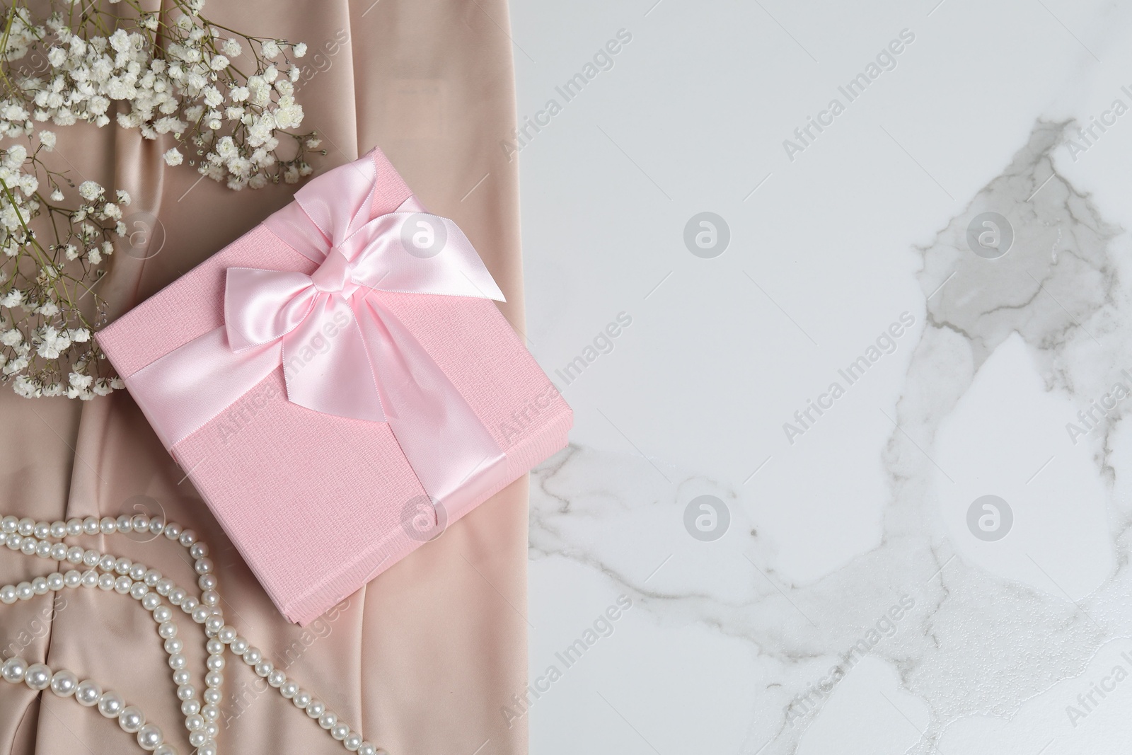 Photo of Beautiful wedding gift, pearl jewelry, silk fabric and gypsophila flowers on white marble table, flat lay. Space for text