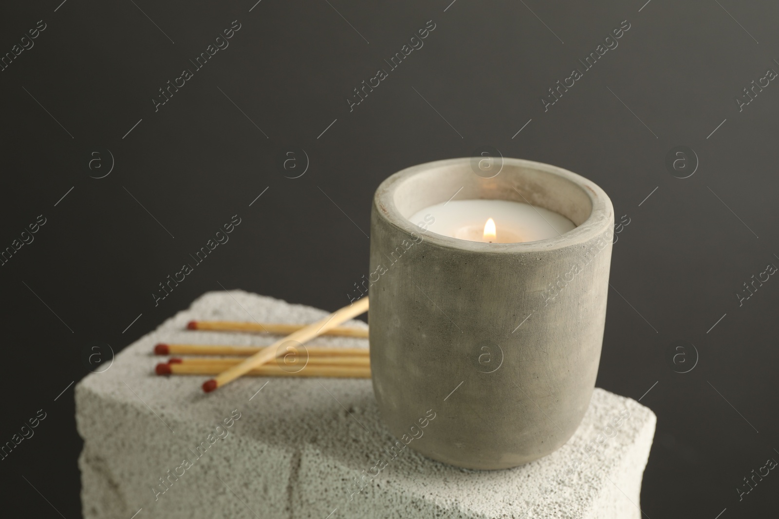 Photo of Burning soy wax candle and matches on concrete block against gray background, space for text