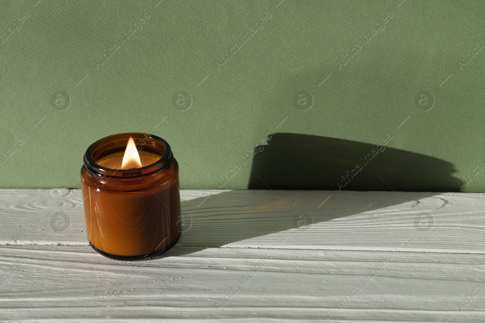 Photo of Burning soy wax candle on white wooden table against green background, space for text