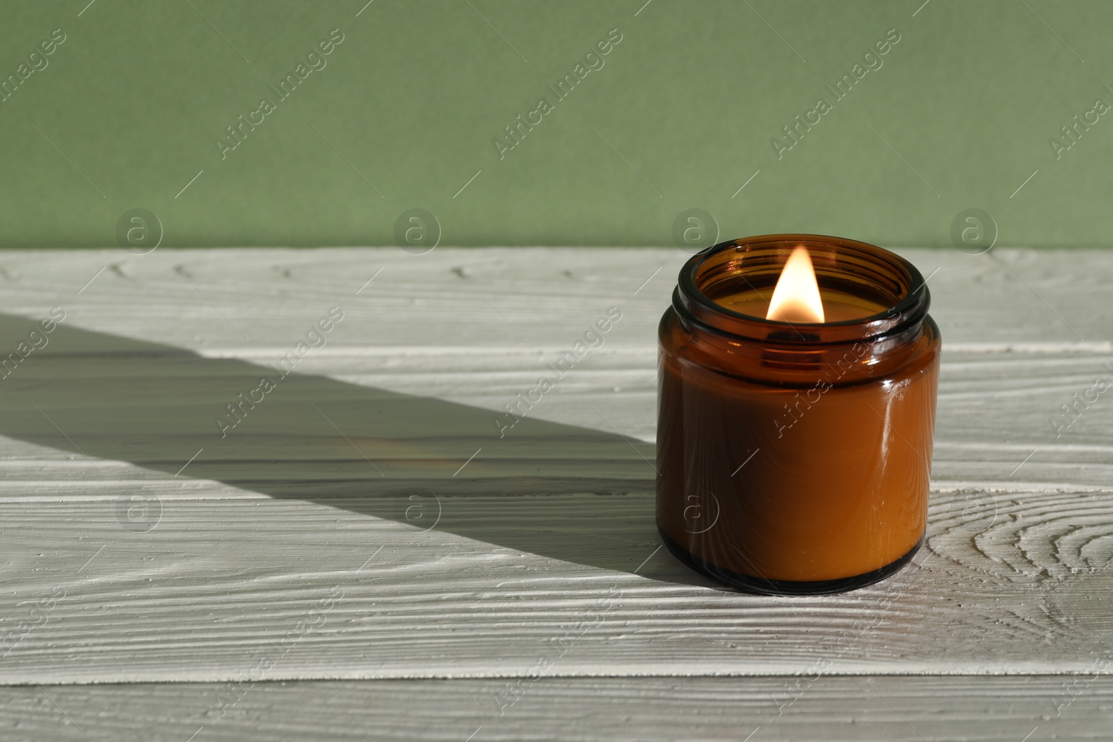 Photo of Burning soy wax candle on white wooden table against green background, space for text