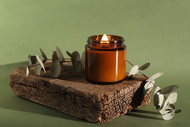 Photo of Burning soy wax candle, decorative piece of wood and eucalyptus branches on green background