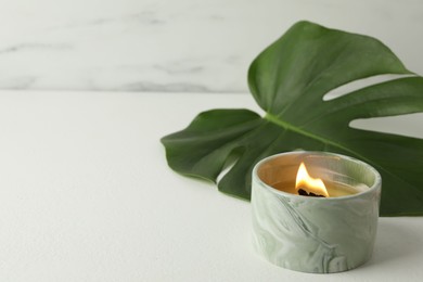 Photo of Burning soy wax candle and monstera leaf on light table, closeup. Space for text