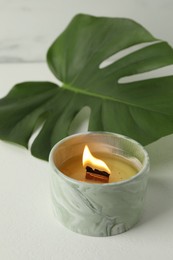 Photo of Burning soy wax candle and monstera leaf on light table, closeup