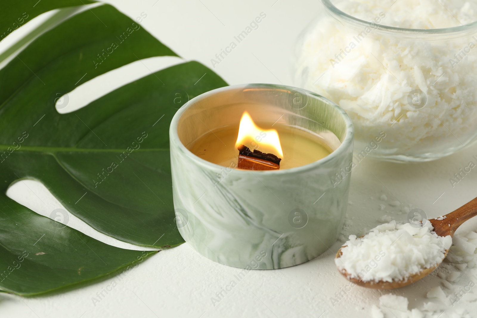 Photo of Burning soy wax candle, monstera leaf and spoon on light table, closeup