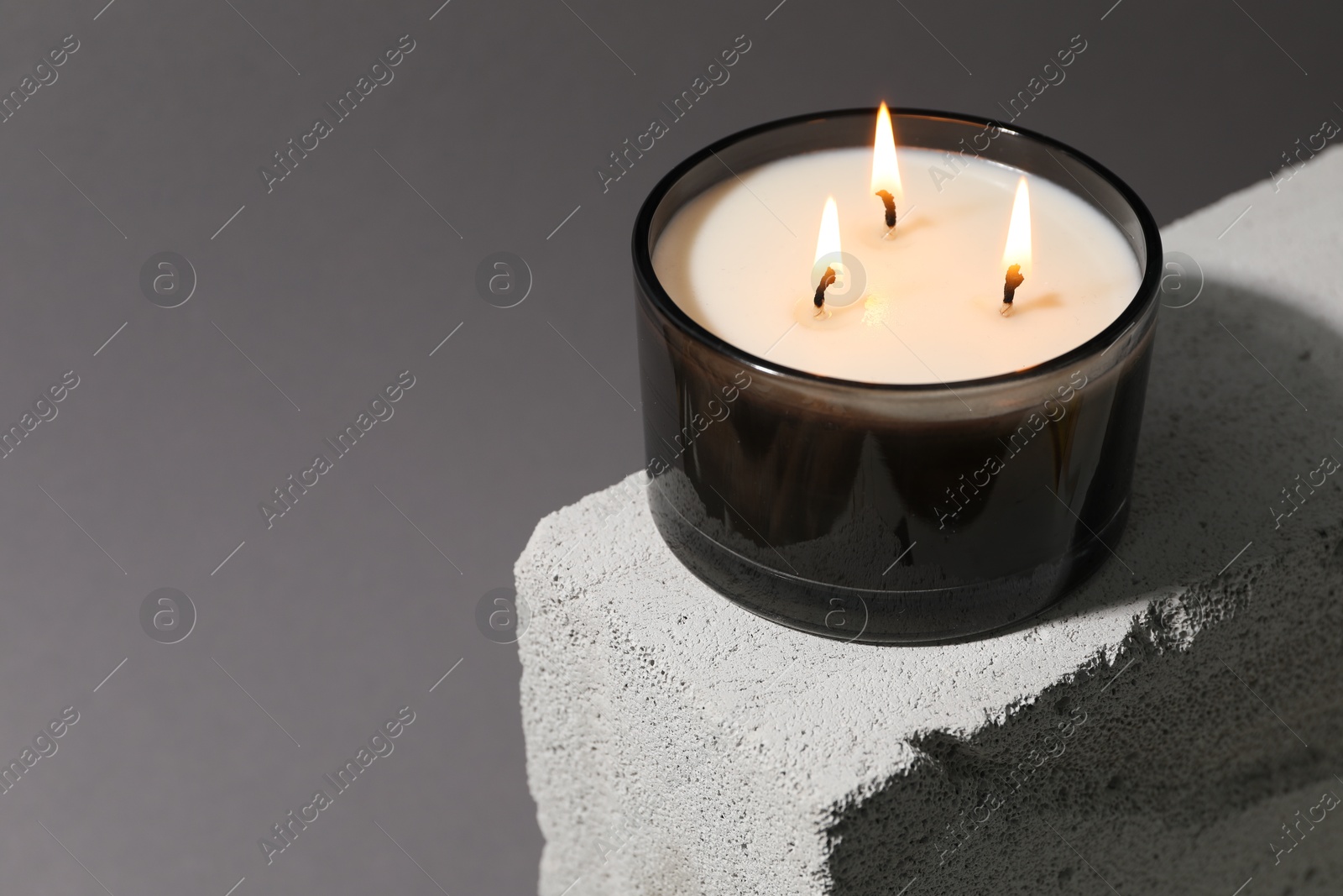 Photo of Burning soy wax candle on concrete block against gray background, space for text